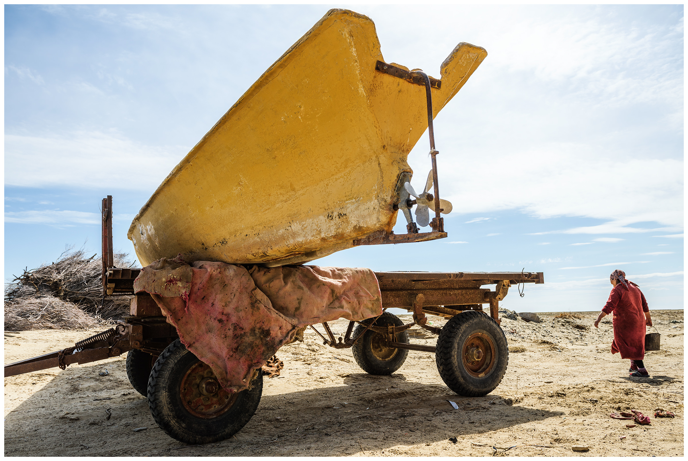 A woman with a bucket in her hand walks past a car trailer loaded with a fishing boat