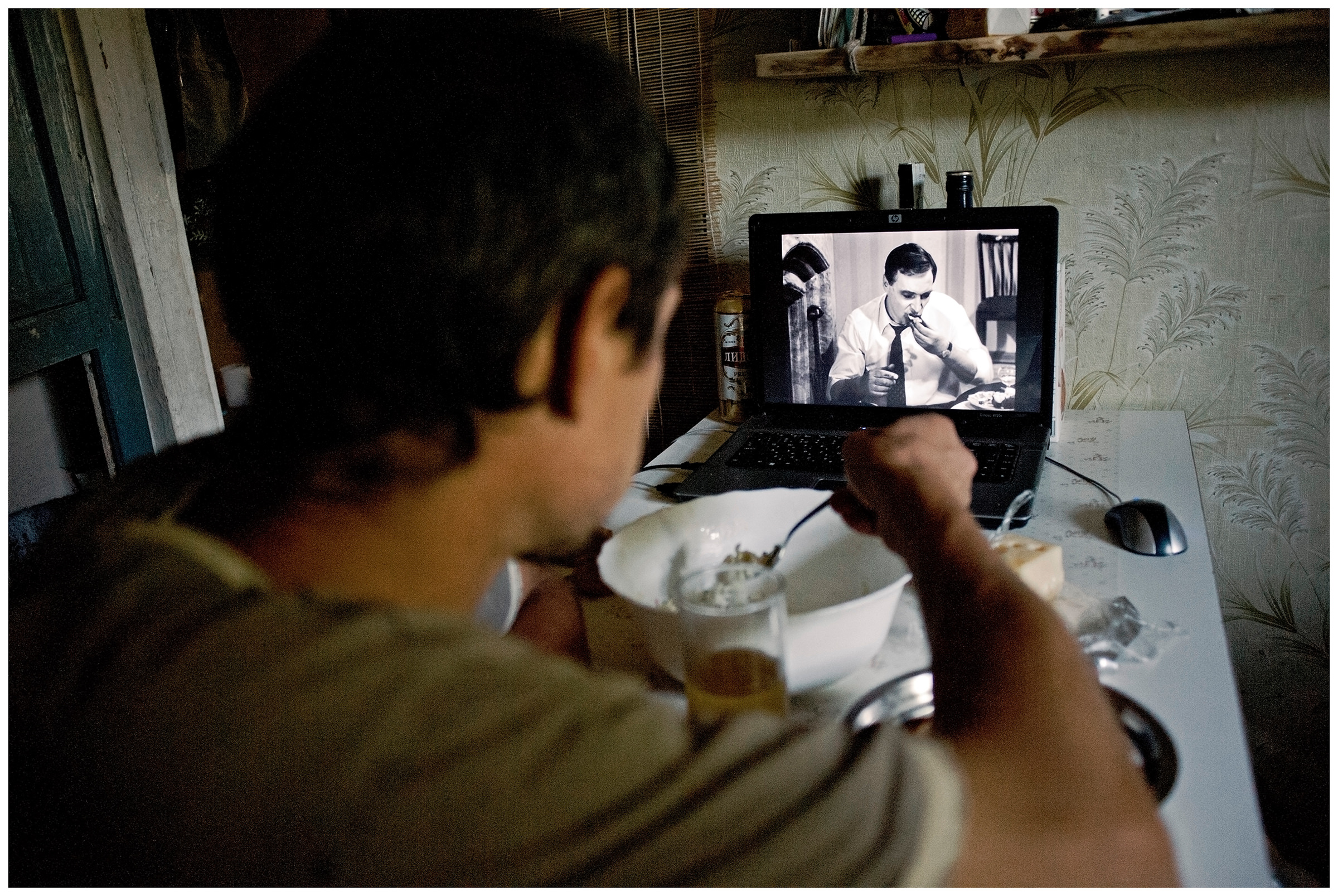 Belarus, village of Chereshlya. A man eats at a table and watches a film on his laptop.