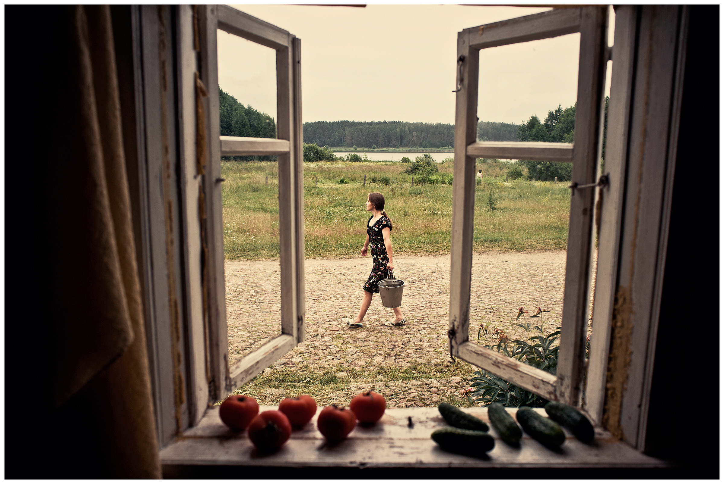 Belarus, village of Chereshlya. A woman with a bucket fetches water. Cucumbers and tomatoes lie on a windowsill.