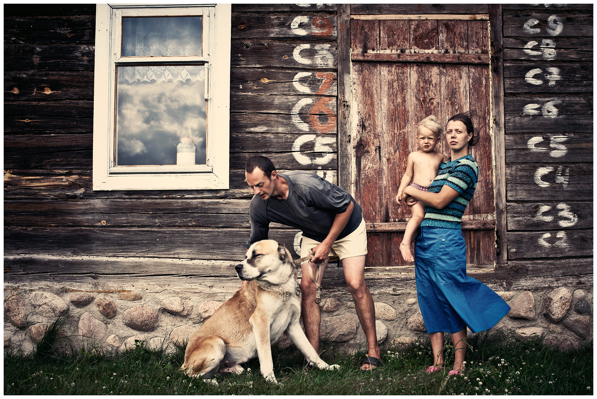 A family with a large shepherd dog stands in front of a wooden house in the village