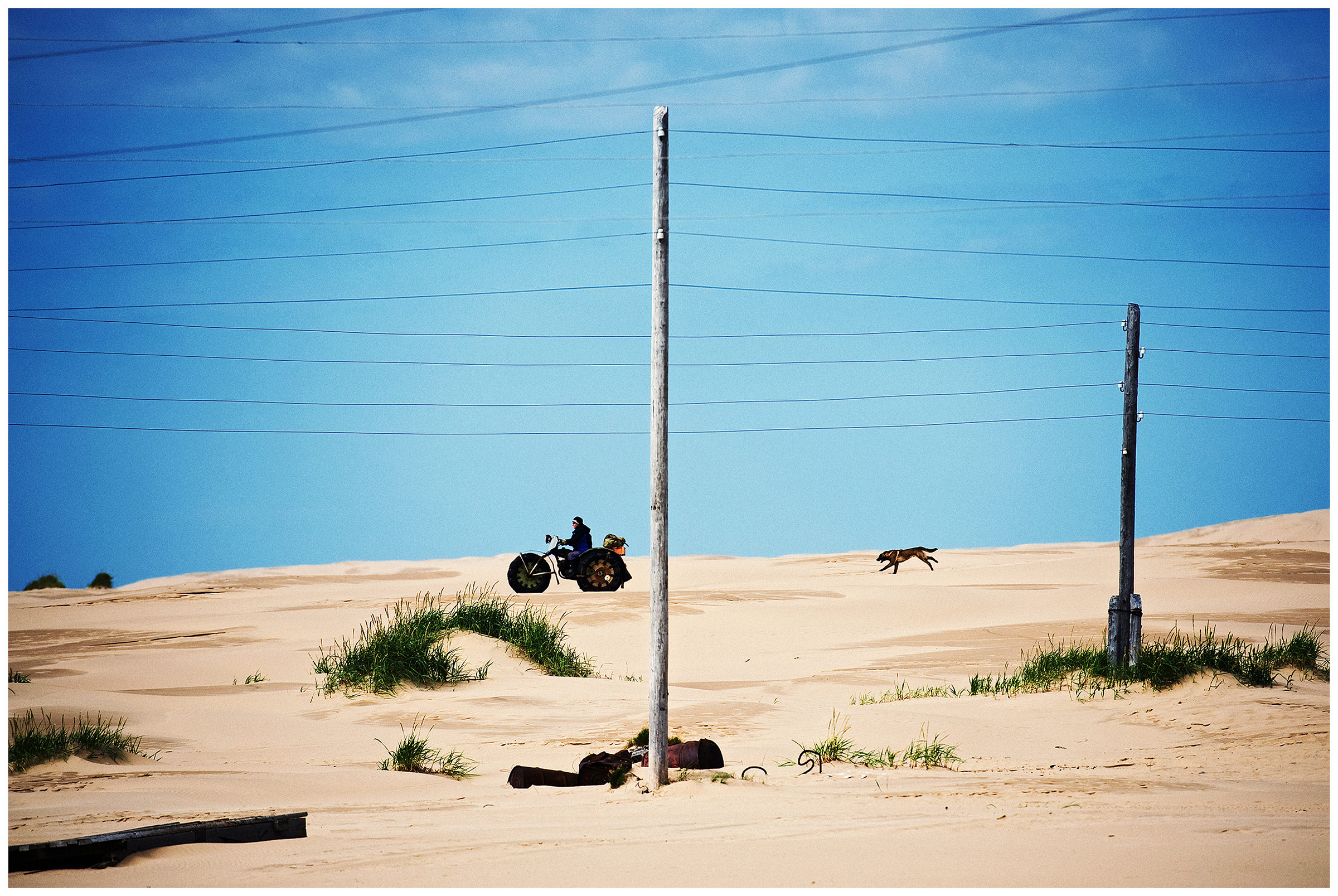 Shoyna village, Russia. A man drives through the sand dunes on a homemade vehicle. A dog follows him.