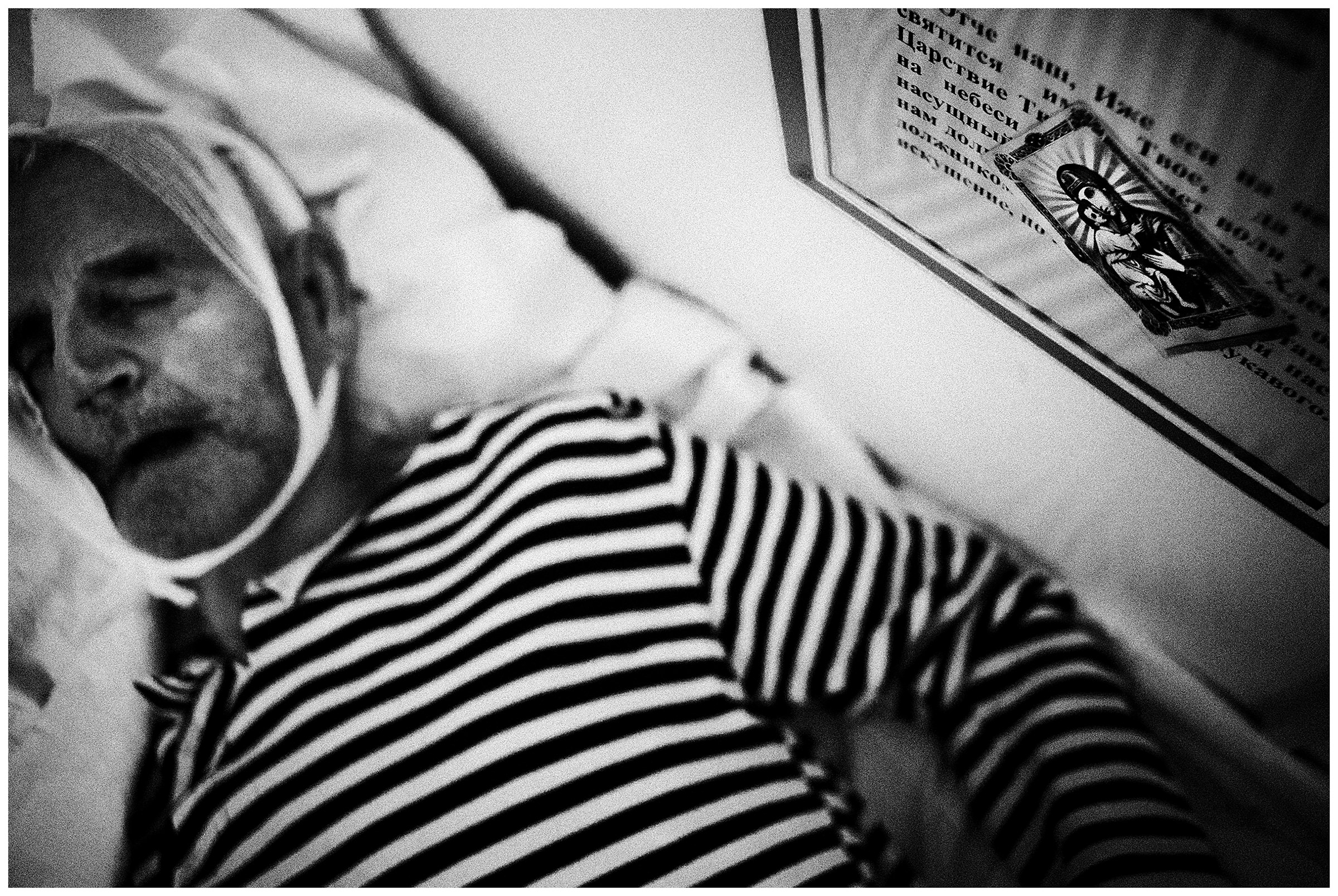 Minsk Hospice, Minsk, Belarus. A patient sleeps under a prayer board over his bed.