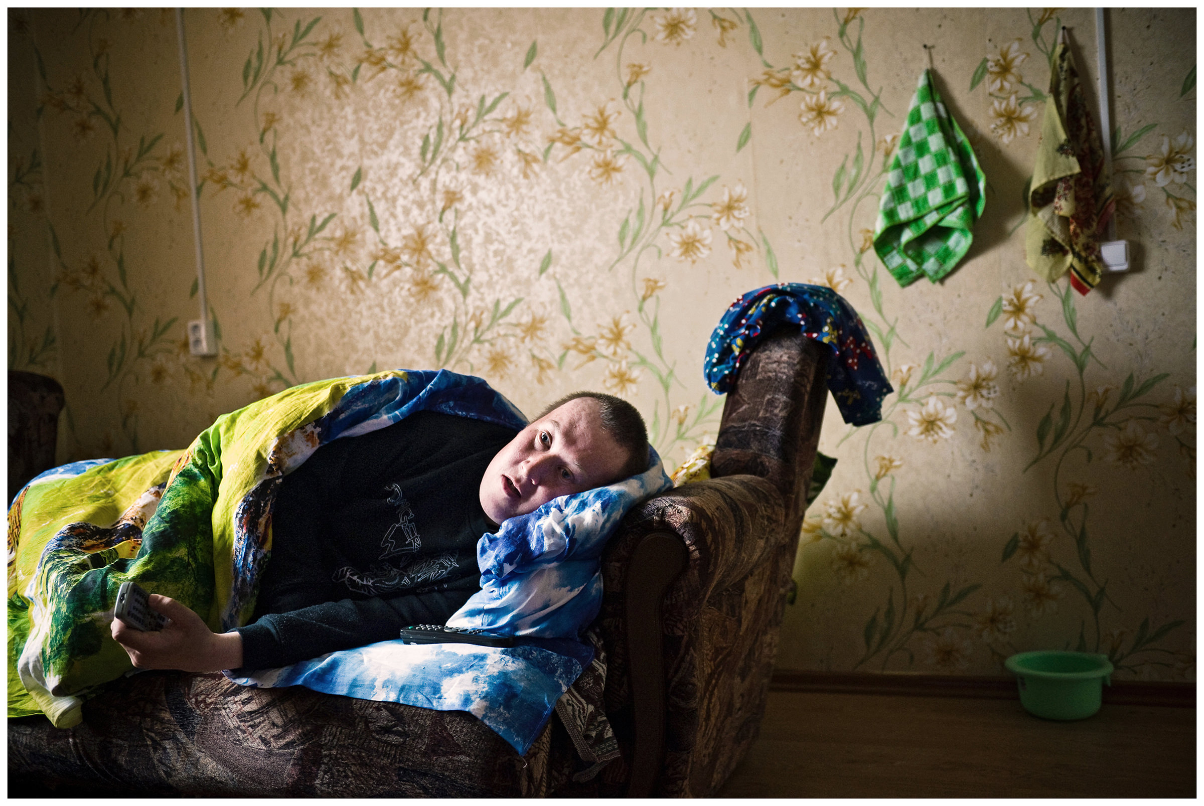 Shoyna village, Russia. A disabled man lies in his bed with a remote control in his hand.