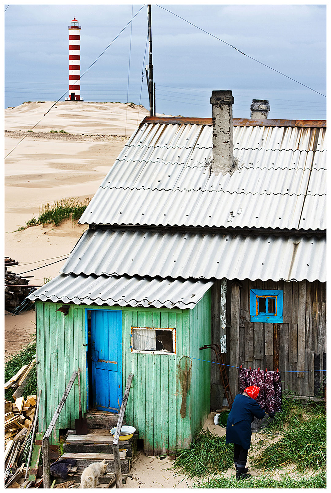 Shoyna village, Russia. A woman in a headscarf hangs laundry on next to her house in front of the lighthouse.