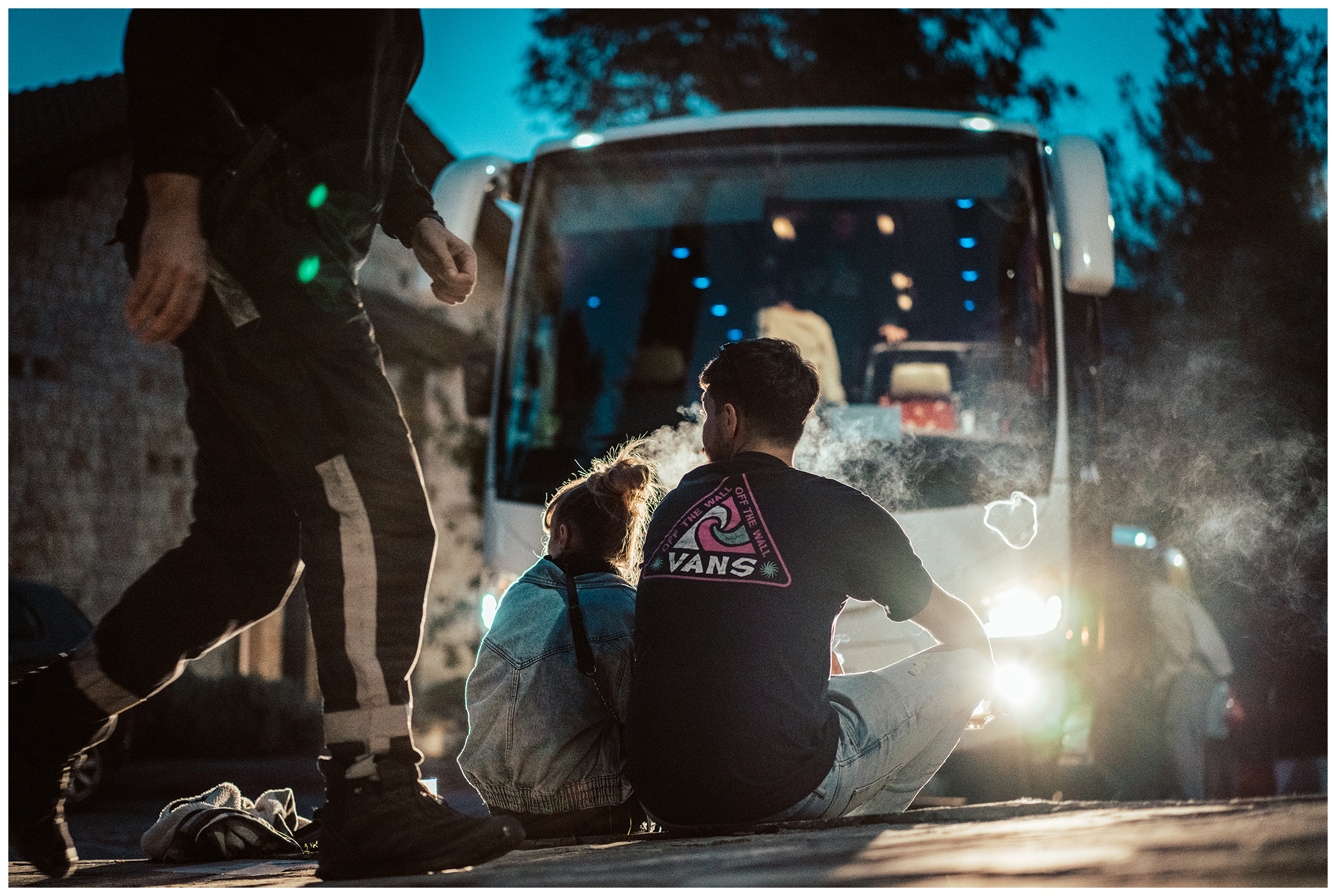Miliou, Cyprus. A man and a woman smoke before boarding a bus that will take them to the airport. An armed police officer walks past them.